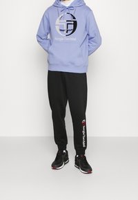 Felpa con cappuccio viola chiaro con grafica del logo nera, abbinata a pantaloni jogger neri che presentano un testo del brand bianco e rosso sulla gamba. Sneakers nere.