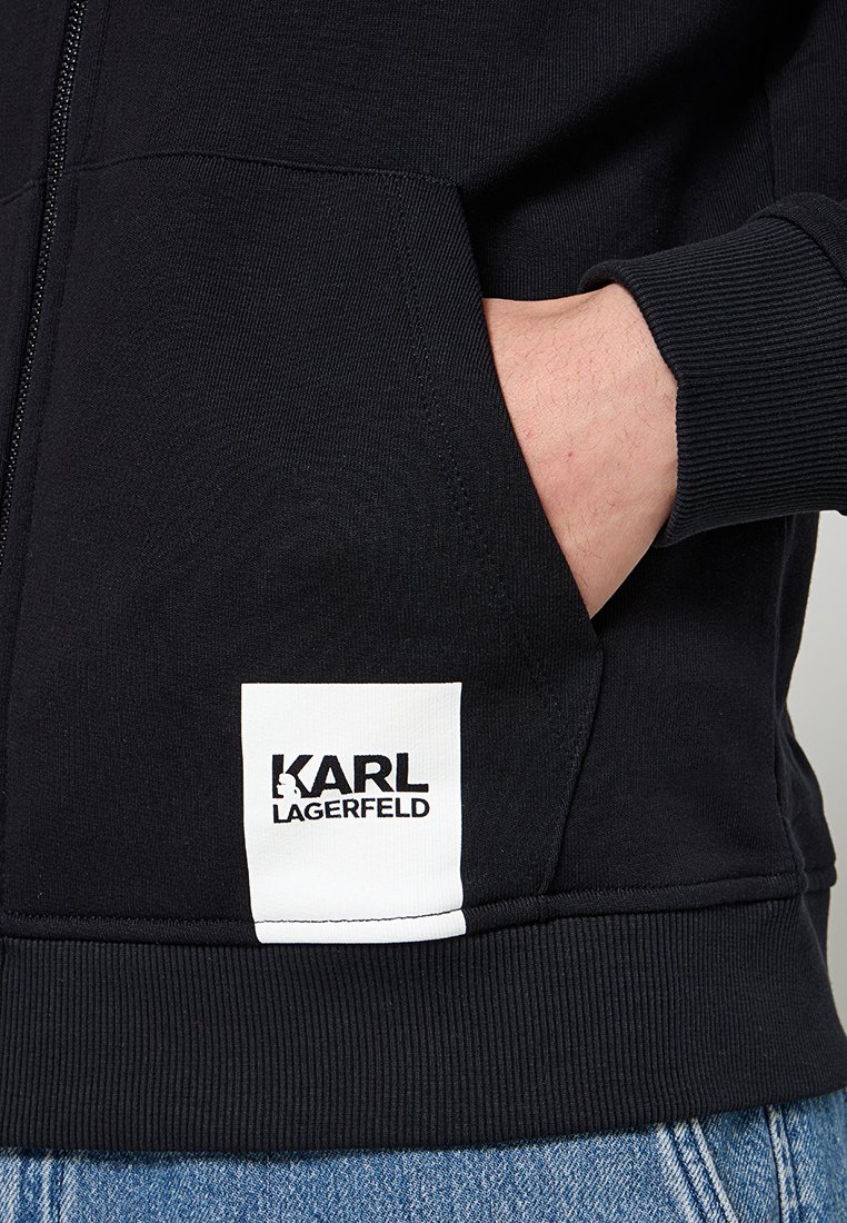 Felpa nera con zip e polsini e orlo a coste. Presenta una tasca frontale e un'etichetta bianca con la scritta "KARL LAGERFELD" in carattere nero.