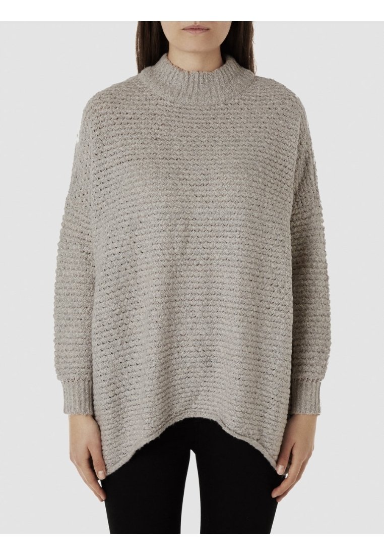 Femme portant un pull en maille ample gris clair texturé avec manches longues et un pantalon noir sur un fond uni.