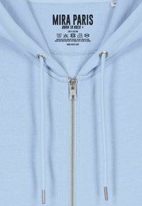 Felpa di cotone azzurro chiaro con zip davanti, accessori in argento, cordini per il cappuccio e un'etichetta con istruzioni per la cura e il marchio.