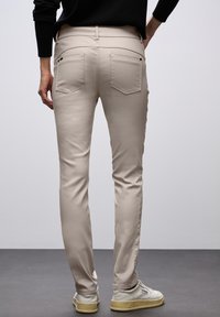 Krémové přiléhavé kalhoty s pěti kapsami, jemnými švy a hladkou texturou, v kombinaci s bílou obuví s robustní podrážkou.