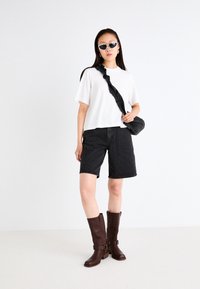 T-shirt blanc à manches courtes, shorts en denim noir, et bottes marron jusqu'aux genoux avec boucles. Sac bandoulière noir et lunettes de soleil blanches complètent le look.