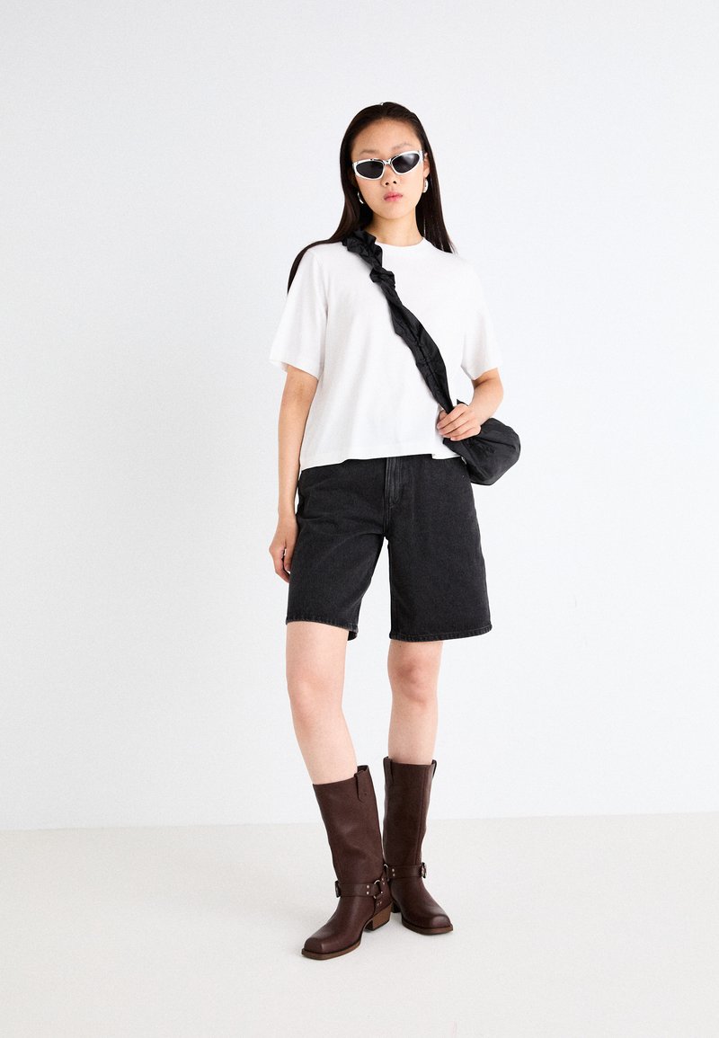 T-shirt blanc à manches courtes, shorts en denim noir, et bottes marron jusqu'aux genoux avec boucles. Sac bandoulière noir et lunettes de soleil blanches complètent le look.