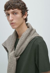 Maglione lavorato a maglia color beige chiaro, disposto su una T-shirt verde scuro; presenta un tessuto morbido e strutturato con una vestibilità ampia e casual.