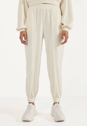 Personne portant un pantalon jogger couleur crème avec ceinture et poignets élastiques, associé à des baskets blanches épaisses.