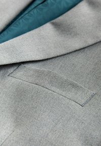 Tela gris texturizada de una chaqueta de traje con solapa con muesca, con un forro interior color verde azulado y un detalle de bolsillo oculto.