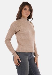 Maglione beige a costine con collo alto, maniche lunghe e un motivo sottile sulle spalle. Abbinato a jeans scuri. Texture morbida.