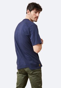 Camiseta de algodón azul marino con cuello redondo clásico, mangas cortas y un pequeño emblema amarillo en el lado inferior.