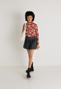 Blusa de mangas longas com flores vermelhas, saia preta plissada e botas de tornozelo pretas. Asegurando uma mala de ombro branca. Fundo neutro.