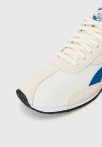Adidași albi cu accente de suede bej, logo albastru, talpă din cauciuc texturată și șireturi plate, având vârful întărit și detalii cusute.