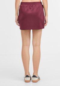 Mini gonna sportiva bordeaux con vita elastica, superficie liscia e design semplice, abbinata a sneaker grigie e nere.