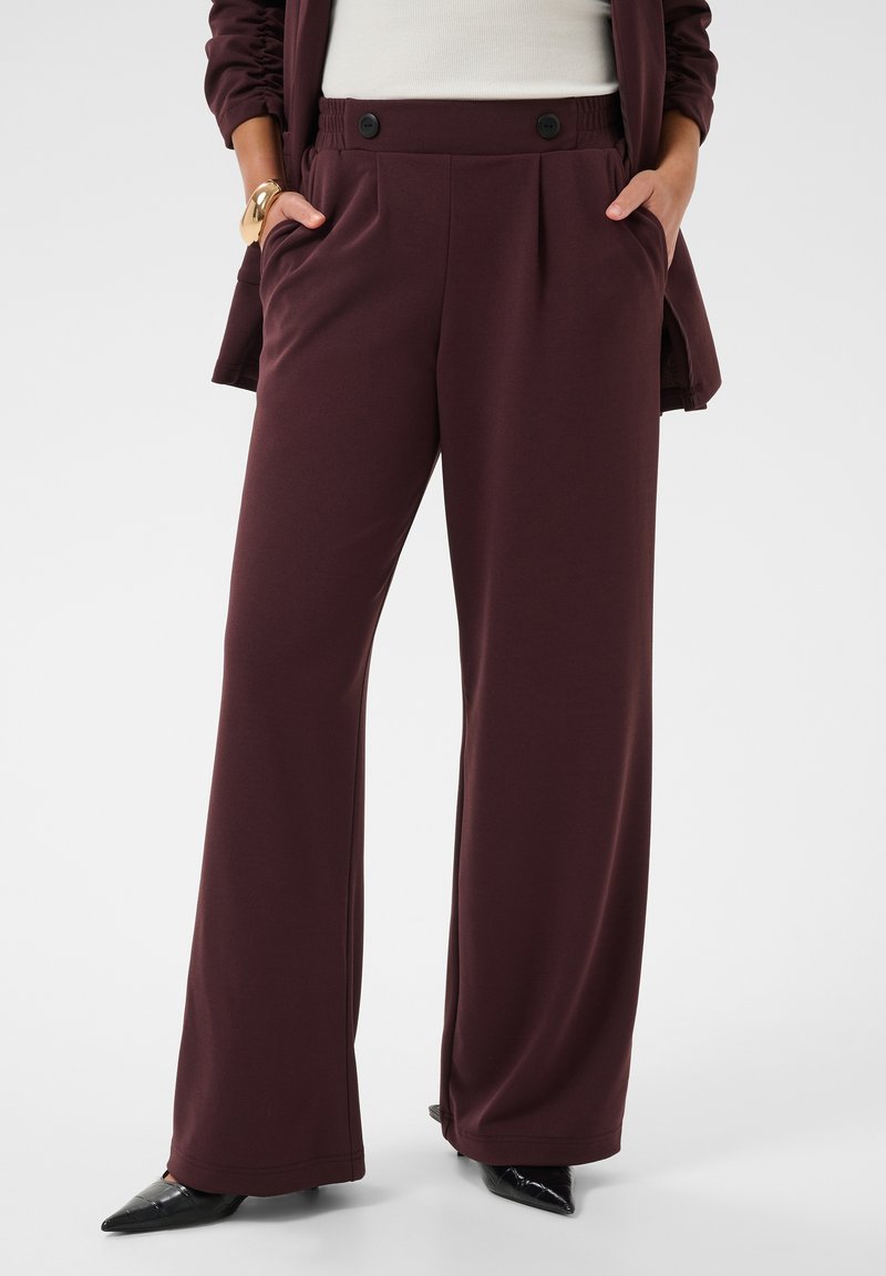 La femme porte un pantalon large violet foncé avec des poches avant, associé à des chaussures pointues noires et un haut blanc partiellement recouvert par une veste assortie.