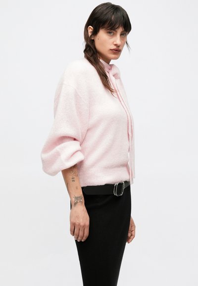 Suéter de punto rosa claro con un ajuste relajado, hombros caídos y dobladillo acanalado. Combinado con una falda negra y un cinturón de cuero.