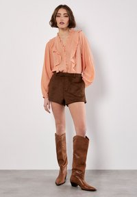 Blusa de melocotón con volantes y cierre de botones, pantalones cortos de ante marrón y botas altas de cuero marrón con punta afilada y tacón bajo.