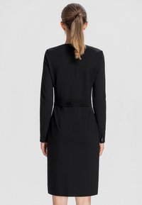 Femme avec une queue de cheval blonde portant une robe noire ajustée à manches longues avec une ceinture, vue de dos sur un fond clair uni.