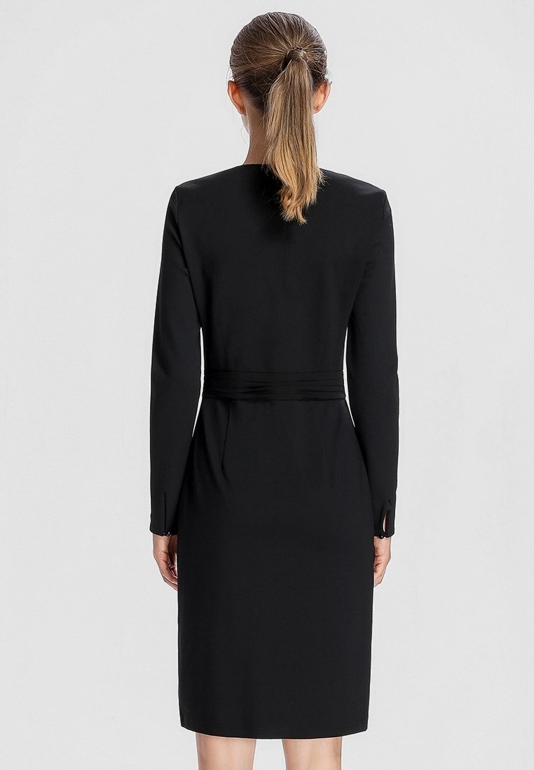 Femme avec une queue de cheval blonde portant une robe noire ajustée à manches longues avec une ceinture, vue de dos sur un fond clair uni.