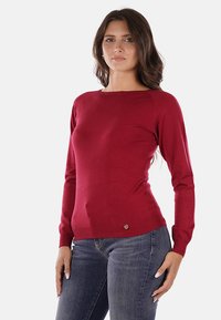 Maglione rosso lavorato a maglia con scollo rotondo, maniche lunghe e design aderente. Presenta una texture liscia e un piccolo logo decorativo all'orlo.