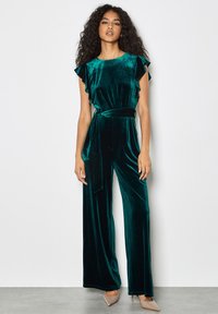 Mono de terciopelo en verde azulado con mangas con volantes, cintura anudada y piernas anchas. Textura suave, diseño elegante y silueta ajustada.