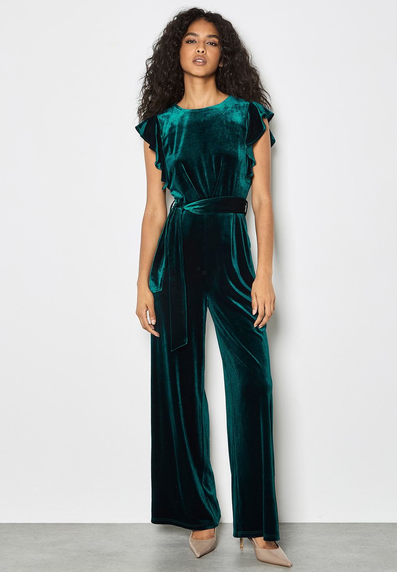 Mono de terciopelo en verde azulado con mangas con volantes, cintura anudada y piernas anchas. Textura suave, diseño elegante y silueta ajustada.