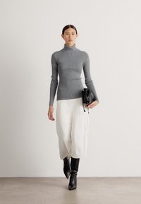 Grauer gerippter Rollkragenpullover, weiße weit geschnittene Hose und schwarze kniehohe Stiefel. Das Model trägt eine schwarze Tasche. Minimalistischer Hintergrund.
