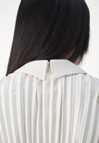 Vue de dos d'une personne aux longs cheveux noirs portant un haut plissé blanc avec un col texturé et une fermeture éclair à l'arrière.