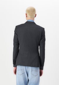 Dunkelgrauer Blazer aus strukturiertem Stoff, taillierte Passform, mit halbseitigem Schlitz hinten und geknöpften Manschetten. Getragen über hellblauen Jeans.
