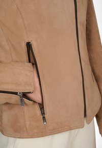 Ljust brunt mockajacka med dragkedjefickor, med en figursydd design och kontrasterande svarta dragkedjor, slät textur och rena linjer.