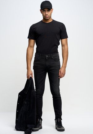 Maglietta nera, jeans skinny neri e giacca di jeans nera tenuta nella mano sinistra. Scarpe nere con dettagli rossi. Sfondo pulito.