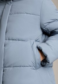 Světlomodrá péřová bunda s prošívanou texturou, vysokým límcem a boční kapsou. Hladký, lesklý materiál s volným střihem.