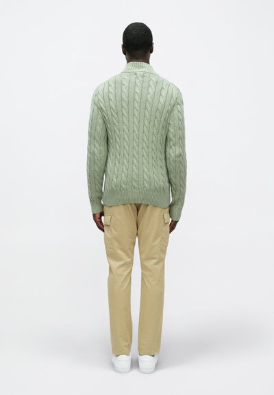 Φωτεινό πράσινο πουλόβερ από πλέξη καλωδίων με ψηλό γιακά, συνδυασμένο με μπεζ cargo παντελόνι και λευκά αθλητικά παπούτσια, βλέποντας από πίσω.