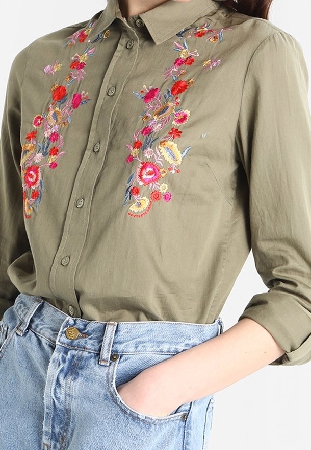 Camicia verde oliva con bottoni e ricamo floreale multicolore abbinata a jeans a vita alta azzurri, mano in tasca.