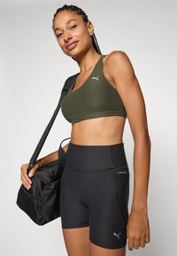 Sostén deportivo de color verde oliva con un diseño de escote en V y logo de PUMA; pantalones cortos negros de cintura alta con una textura suave y detalles de PUMA; llevando un bolso negro.