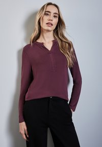 Langärmliges, kastanienbraunes Oberteil mit V-Ausschnitt, weicher Textur und dezenten Nähdetails, kombiniert mit schwarzen Hosen.