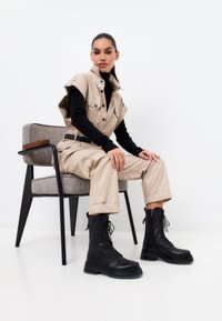 Mono utility beige de mangas cortas, con un cuello de tortuga negro debajo y botas de combate negras con cordones; sentado en una silla negra y gris.