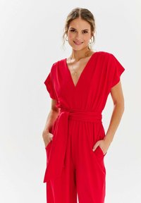 Czerwony kombinezon z głębokim dekoltem w kształcie V, krótkimi rękawami i przewiązanym paskiem w talii. Posiada boczne kieszenie i gładki, lekki materiał.