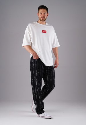 Weißes oversized T-Shirt mit rotem Etikett, kombiniert mit schwarzen gemusterten Hosen mit vertikalen Streifen; abgerundet mit weißen Sneakers.