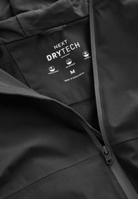 Crna jakna s vodootpornim dizajnom, mekanom unutrašnjom podstavom, zavarenim šavovima i prednjim patentnim zatvaračem. Na etiketii je oznaka "NEXT DRYTECH" i veličina M.