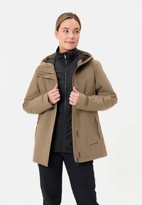 Bruine waterdichte jas met capuchon, voorzien van een zwarte binnenvoering. Heeft ritssluitingen en een subtiel logo aan de onderkant.