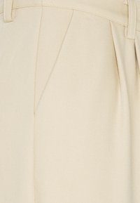 Pantalons taillés beiges en tissu texturé, avec un devant plat, des plis et des passants de ceinture. Détails de la couture visibles.