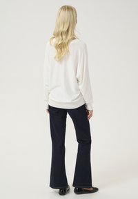 Balta pārāk liela džempera ar ribotu apakšmalu, kombinācijā ar tumši zilām platām biksēm un melniem apaviem, redzams no aizmugures. Gluda tekstūra.