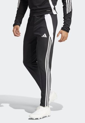 Mann trägt schwarze Adidas-Trainingshose mit weißen Seitenstreifen und Logo, kombiniert mit weißen Fußballschuhen, steht vor einem schlichten Hintergrund.