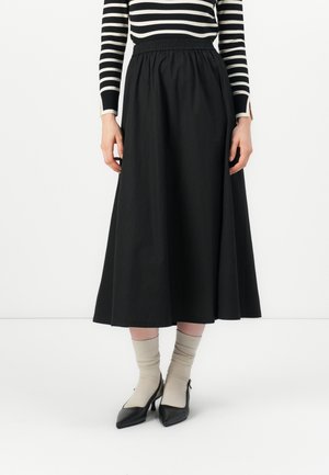 Jupe noire longueur cheville en tissu lisse avec une taille élastique. Portée avec des chaussettes beiges et des escarpins noires à bout pointu.