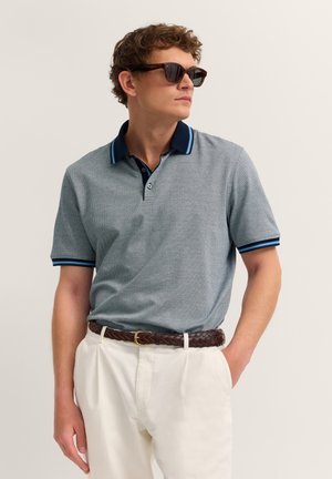 Mann mit Sonnenbrille, blau gemustertes Polohemd mit navyfarbenem Kragen, weiße Hose und brauner geflochtener Gürtel, steht mit einer Hand in der Tasche.