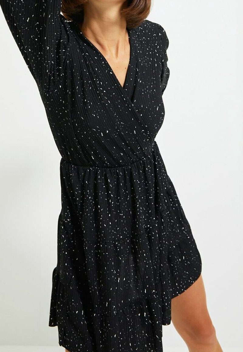 Femme portant une robe noire à manches longues avec des taches blanches et un décolleté en V cache-cœur sur un fond clair uni.