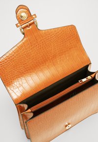 Beżowa torebka z tłoczoną skórą w wzór krokodyla z zapięciem na złote okucia. Wnętrze wyściełane czarną podszewką z zamykaną na suwak kieszenią. Otwarty fason.