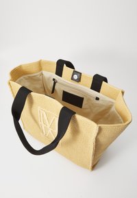 Geel geweven tas met zwarte stoffen handvatten. De open bovenkant onthult een beige interieur, met een ritsvak en een patchdetail aan de binnenkant.