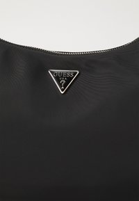 Borsa nera in nylon con un logo metallico triangolare recante il testo "GUESS" e "U.S.A." Chiusura con zip nella parte superiore con una texture liscia.