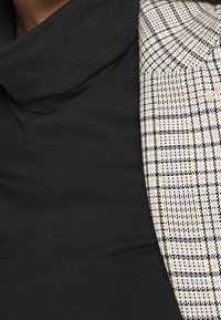 Maglione a collo alto nero in tessuto morbido, abbinato a un blazer stampato con un disegno a scacchi bianco, nero e giallo.