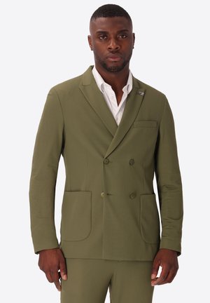 Uomo che indossa una giacca a doppio petto verde oliva con tasche applicate e camicia bianca, in piedi su sfondo bianco.