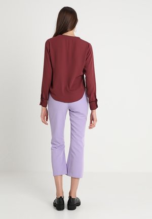 Frau mit langem braunem Haar, die ein burgunderrotes Langarmshirt, fliederfarbene knöchellange Hose und schwarze Schuhe trägt und vor einem schlichten weißen Hintergrund steht.